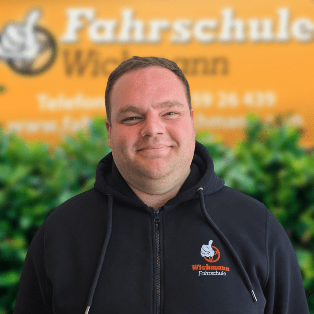 Gunnar, Fahrlehrer der Fahrschule Wichmann Delmenhorst, lächelt vor dem gelben Firmenschild mit Logo und T-Shirt.