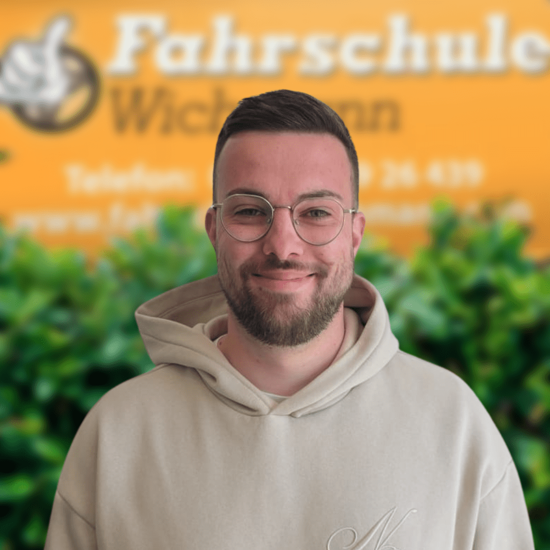 Fahrlehrer der Fahrschule Wichmann in schwarzem Logo-Pullover vor gelbem Hintergrund mit grünem Buschwerk.