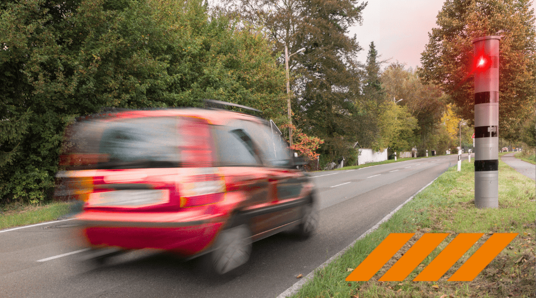 Blurred red car speeding along a rural two-lane road with a red warning light on a striped post and orange roadside hazard stripes nearby.